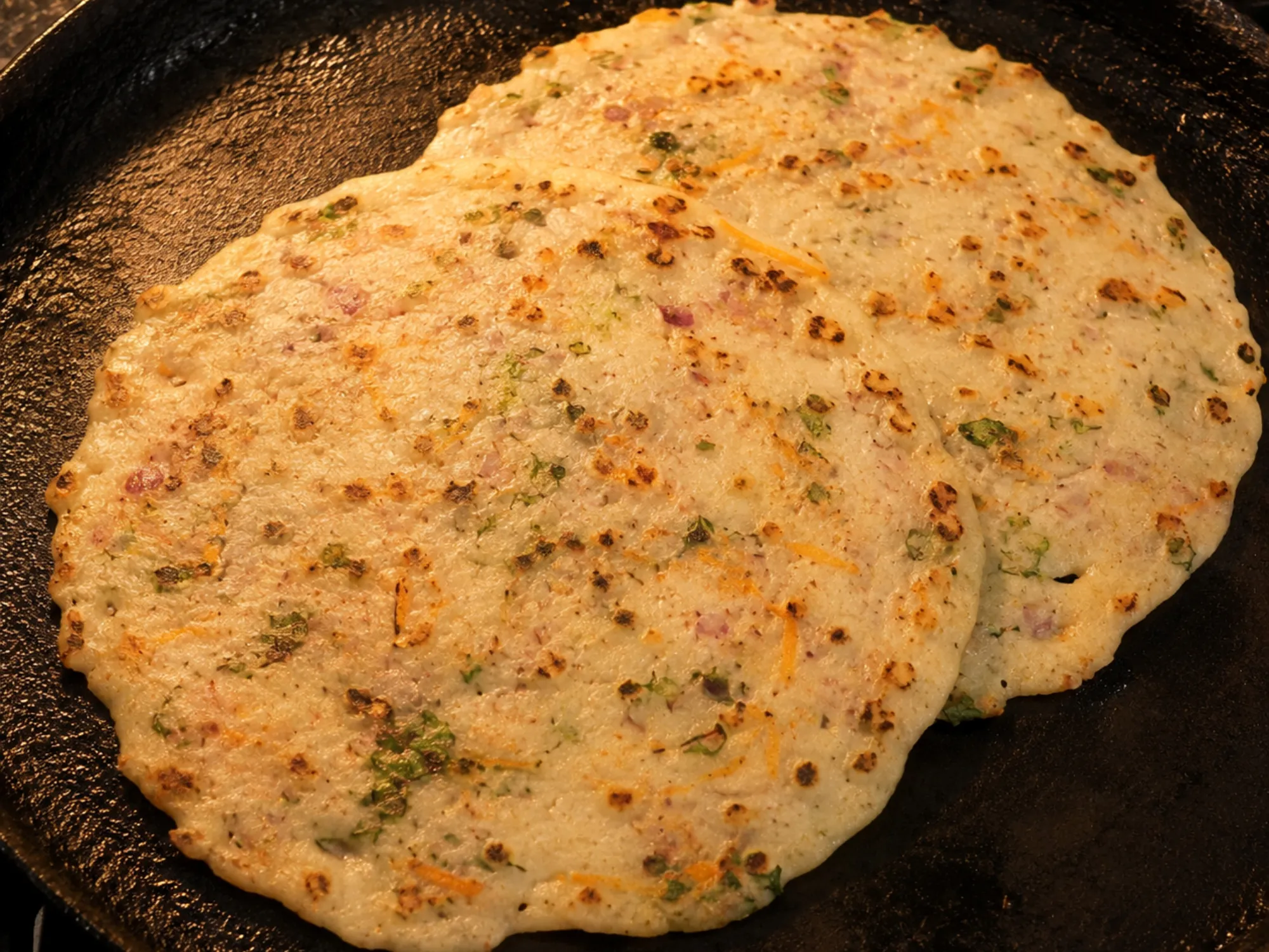 A thin white rice rotti on a tawa with brown spots, flecked with onion, coriander and grated carrot