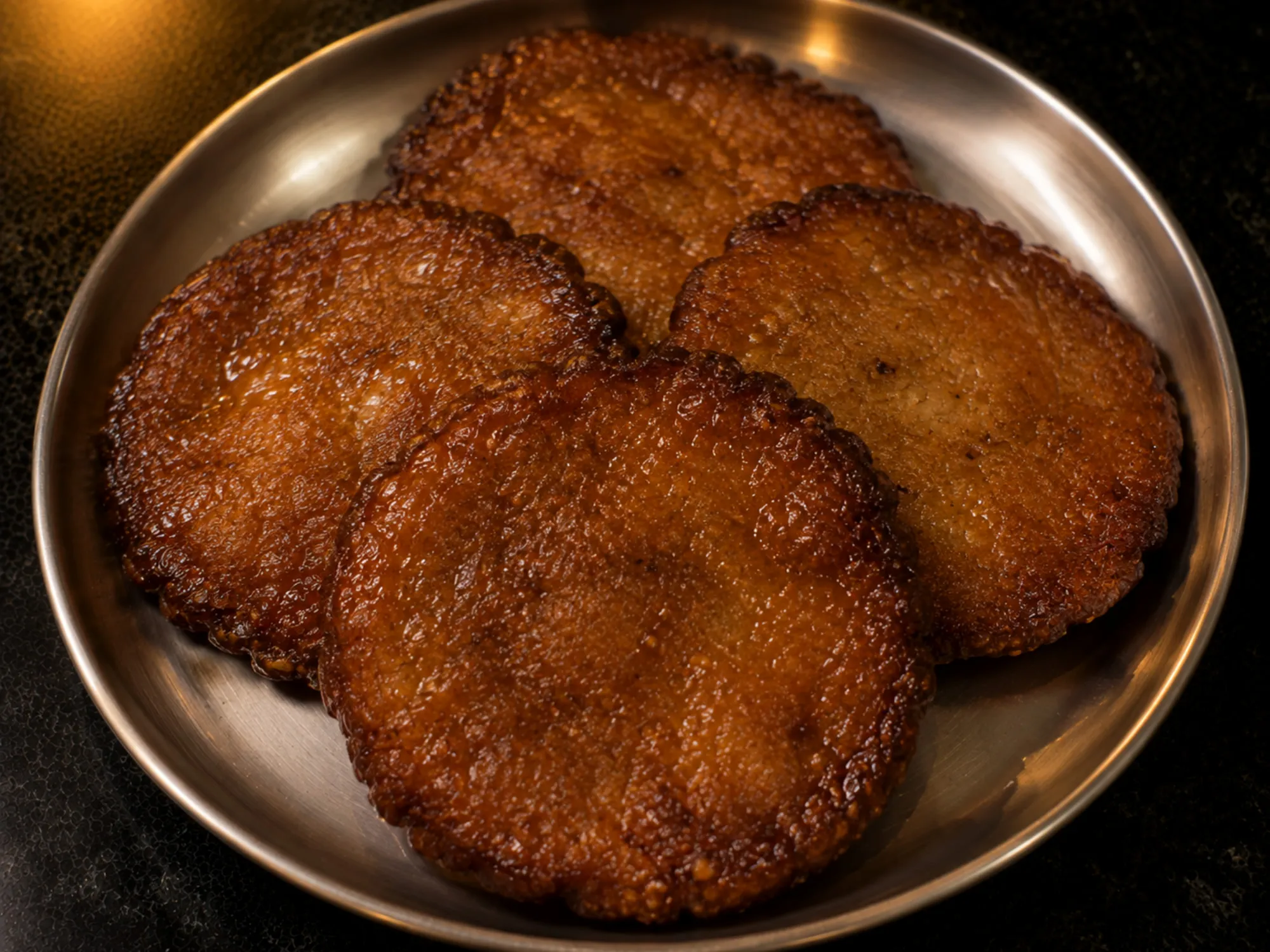 Four dark reddish-brown adhirasam stacked on a steel plate, edges slightly rippled