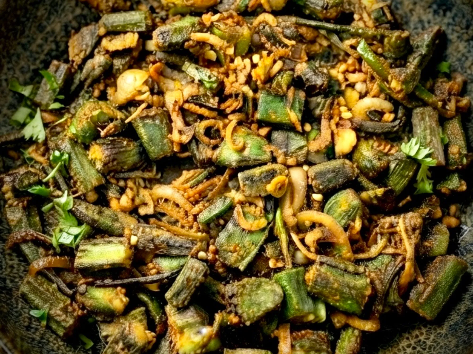 A kadai of dry okra fry, pieces browned at the cut edges and coated in red-brown masala, scattered with coriander